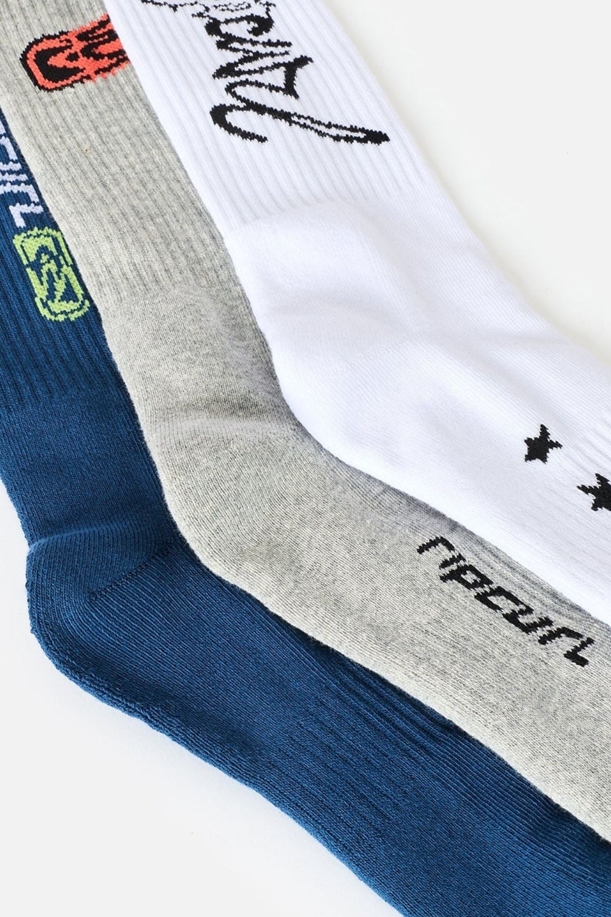 Three pairs of socks from the Rip Curl Retro Crew Sock 3 Pack—one navy blue, one gray, and one white—are laid side by side. Each pair features black or colorful text and retro-inspired graphic designs near the top, along with cushioned comfort and supportive arch construction.