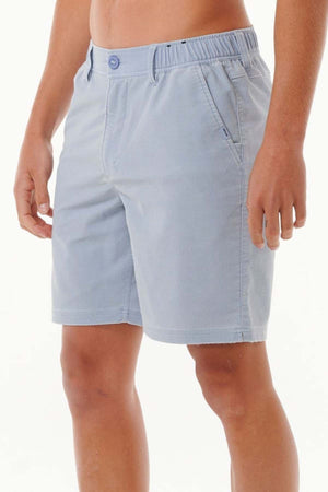 A person stands against a plain background wearing Rip Curl Saltwater Culture Cord Boardwalk Shorts in light blue. These shorts feature quick-dry technology, an elastic waistband with button closure, and side pockets. They end above the knee. The individual is shirtless and only partially visible from the chest down.