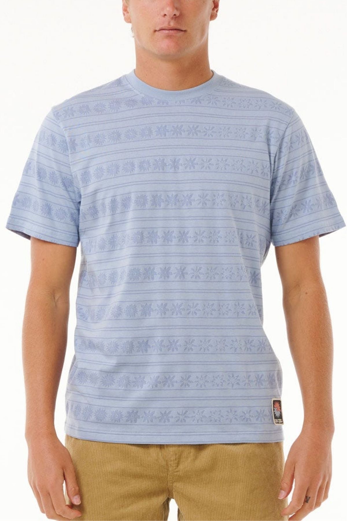 A person wears the Rip Curl Saltwater Culture Stripey Tee in light blue with a subtle jacquard stripe pattern and tan pants, standing against a plain white background. Their face is partially out of frame.