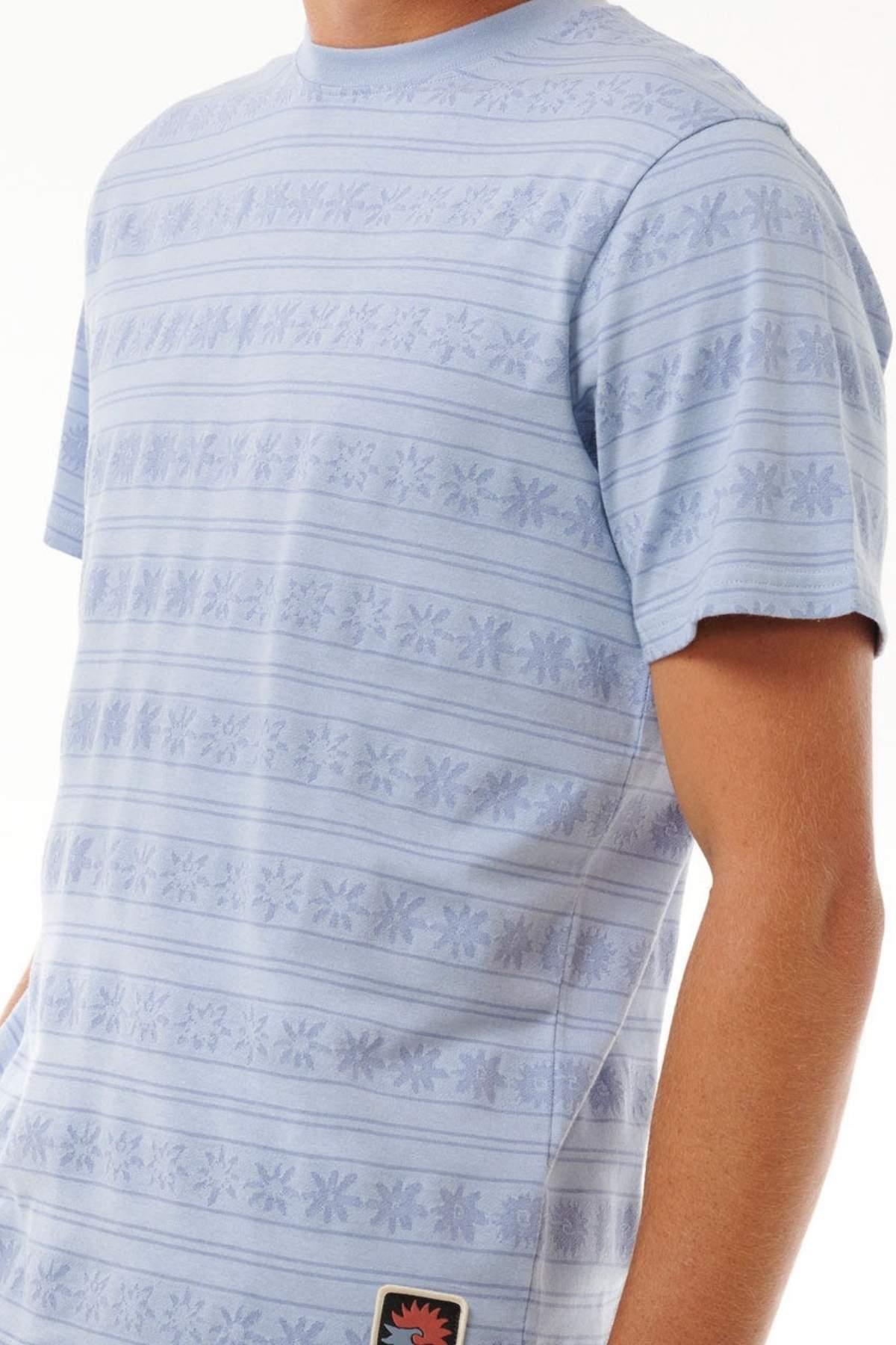 A person wears the Rip Curl Saltwater Culture Stripey Tee, a relaxed fit light blue top with jacquard stripe and floral patterns, made from 100% cotton, shown from shoulders to waist against a plain white background.