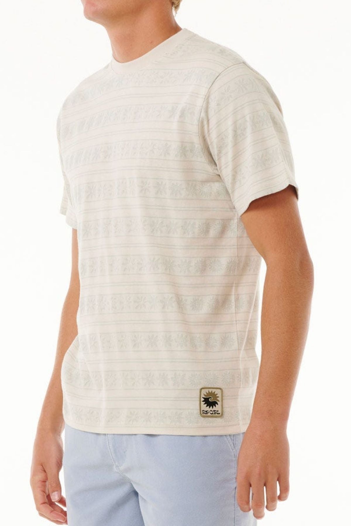 A person dressed in a Rip Curl Saltwater Culture Stripey Tee with light blue pants stands against a plain background. The tee has a small design patch near the hem, capturing the Saltwater Culture vibe. The person's face is not visible.