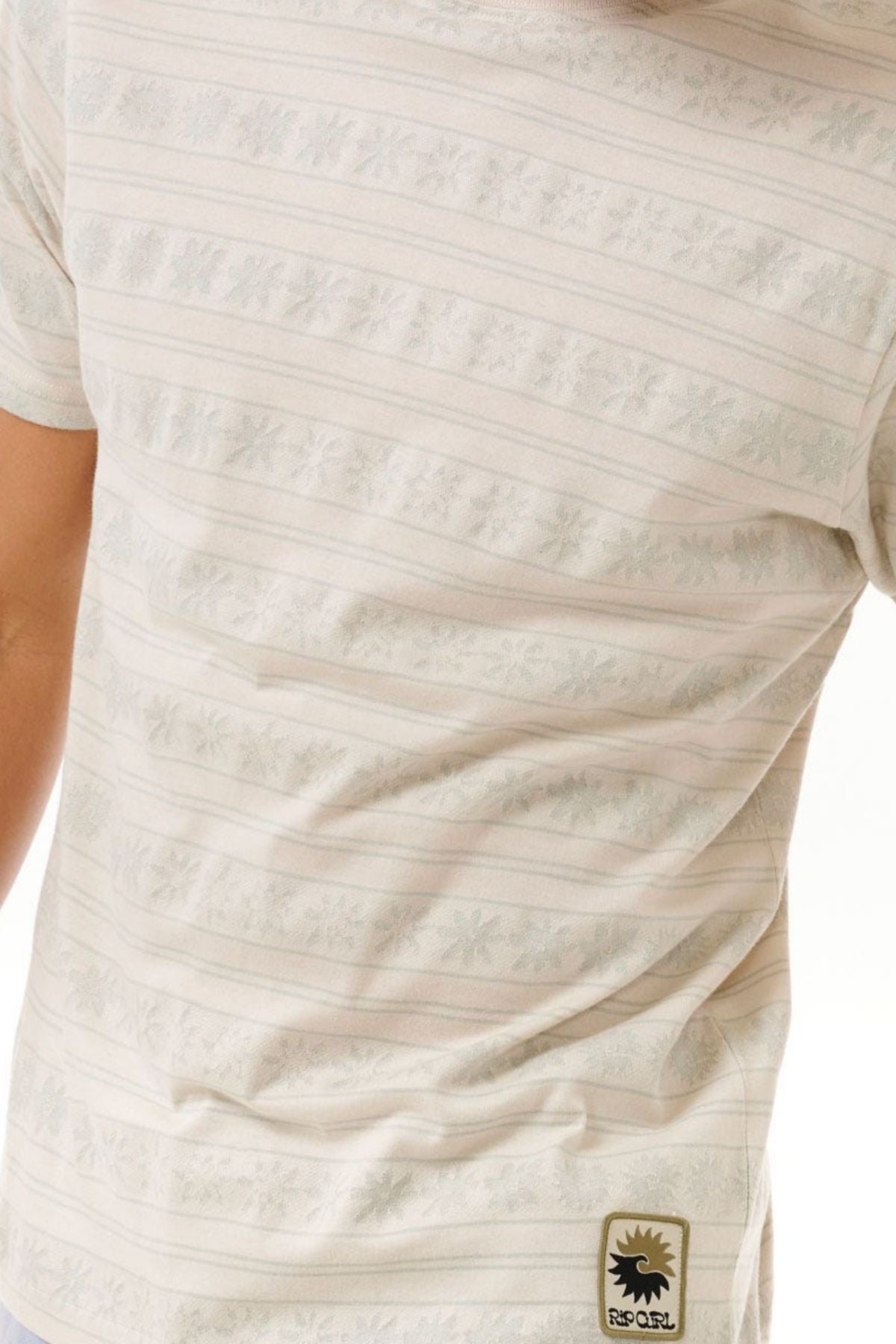 A person is wearing the Rip Curl Saltwater Culture Stripey Tee, featuring subtle jacquard stripes and a small logo at the bottom. This light-colored shirt is short-sleeved with a snug fit.