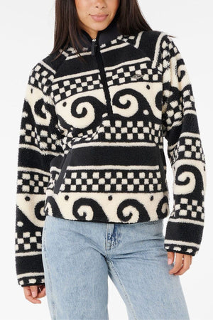 A woman wears the Rip Curl San Carlos Polar Fleece, a fuzzy black and white pullover with bold swirl and checkered patterns, half-zip front, and light blue jeans. The background is plain white.