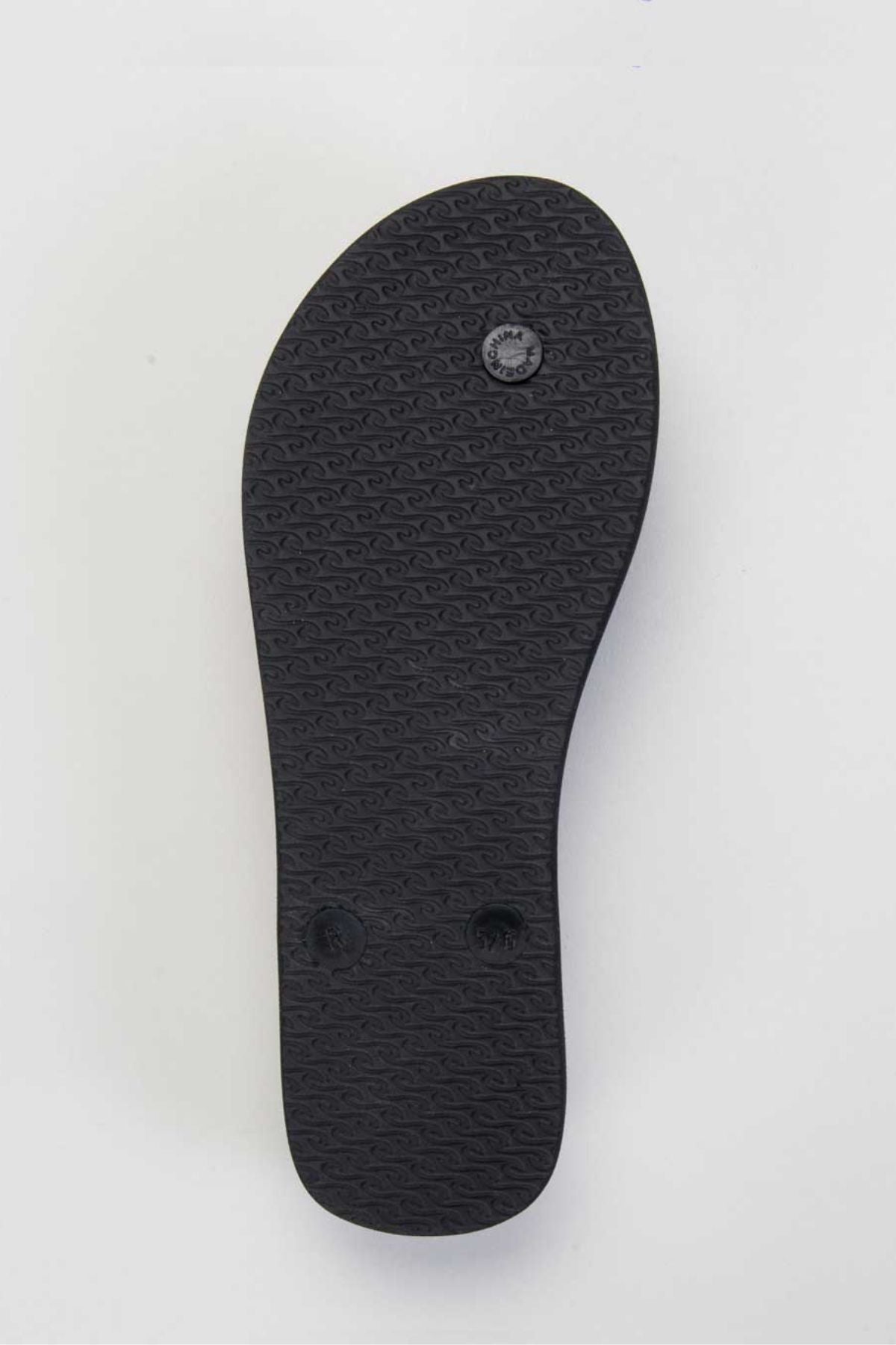 The image showcases the sole of a black sandal made from BLOOM™ algae blended foam EVA, featuring a textured pattern with small, repeating wave-like designs similar to those on Rip Curl thongs. The design includes two circular indentations near the heel and a smaller circular mark nearer the center. This pattern is characteristic of the Rip Curl Script Wave Bloom Thongs.