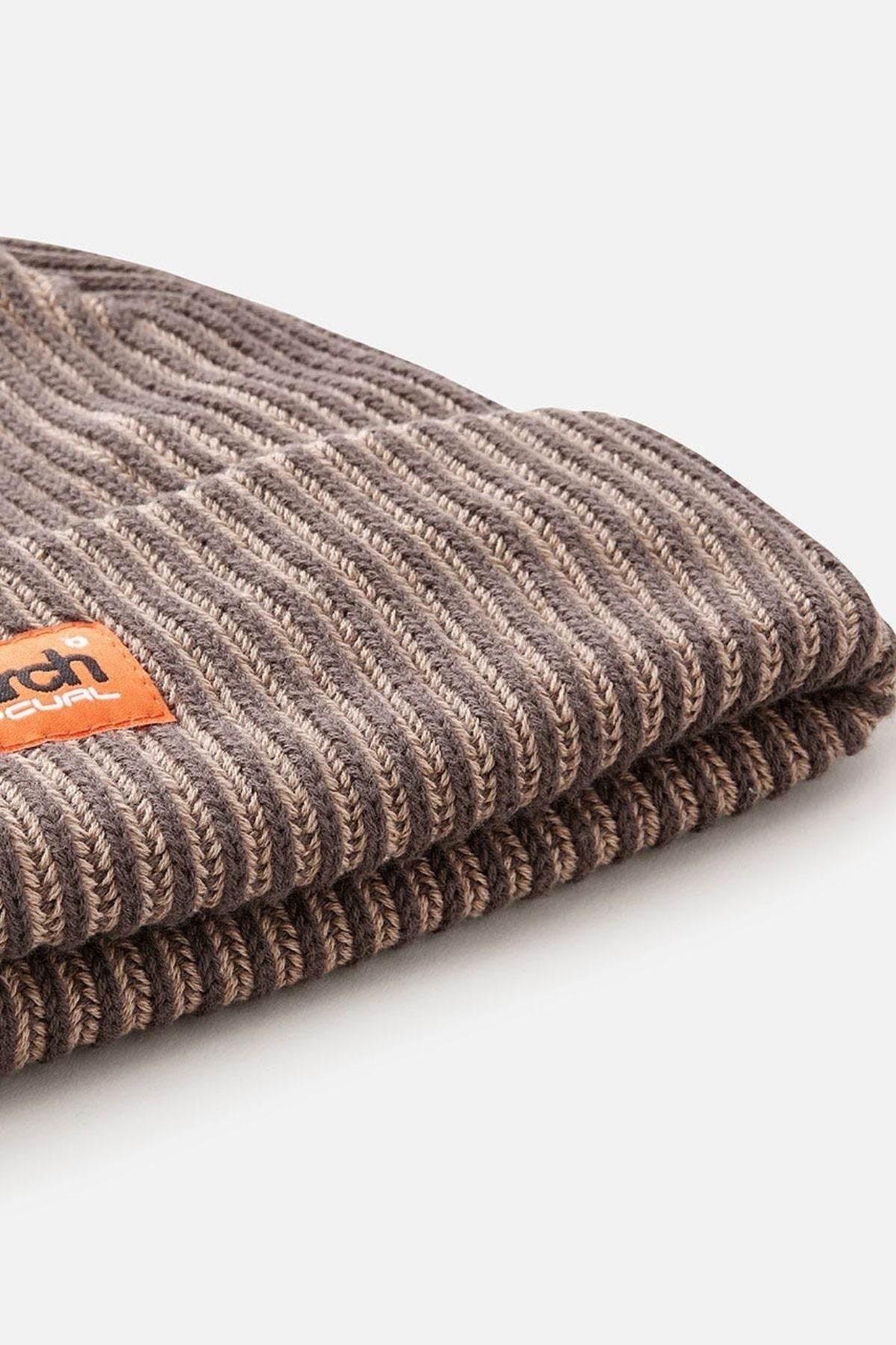 Close-up of the Rip Curl Search Chunky Reg Beanie in brown ribbed knit with a folded brim and a small leather patch showing embossed Rip Curl text, photographed on a plain white background.