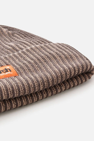Close-up of the Rip Curl Search Chunky Reg Beanie in brown ribbed knit with a folded brim and a small leather patch showing embossed Rip Curl text, photographed on a plain white background.