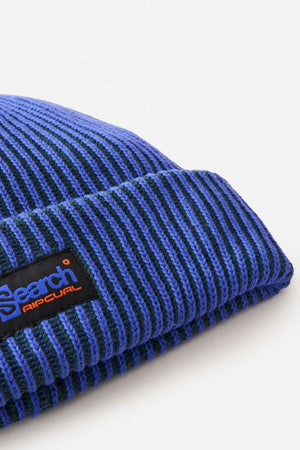 Close-up of the Rip Curl Search Chunky Reg Beanie in blue and black ribbed knit, made from 100% cotton. Features a folded brim and a black patch label with "Search Rip Curl" text—ideal for cold weather.