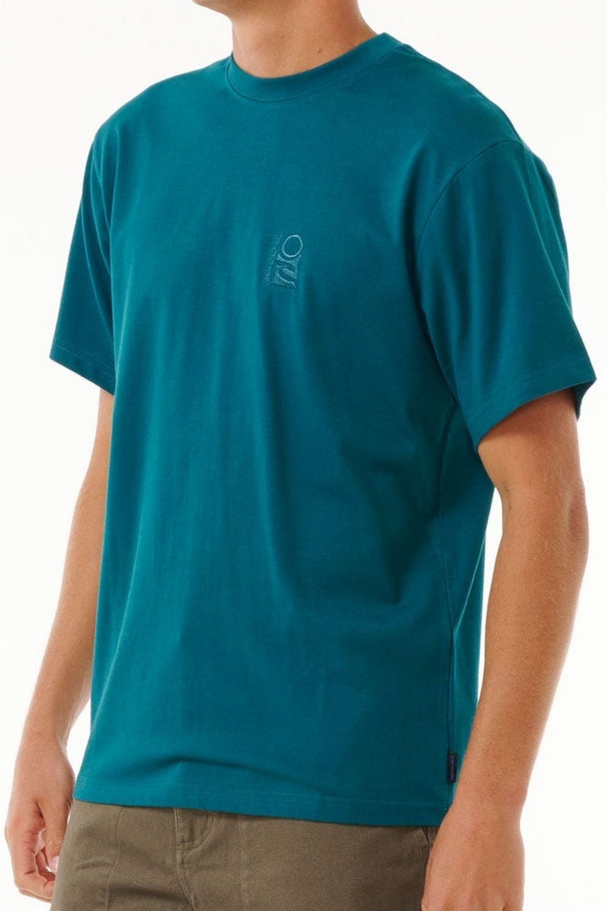 A person is wearing a teal Rip Curl Search Embroidery Tee featuring a small vertical logo on the chest, paired with olive green cotton pants, set against a plain white background.