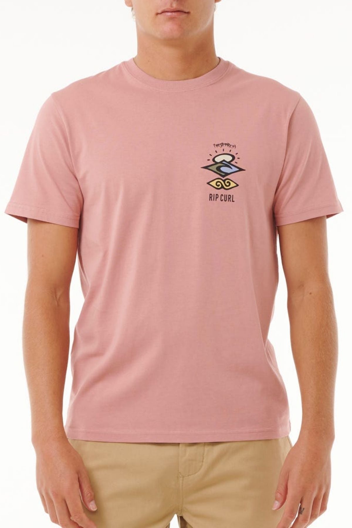 A person wearing a light pink Rip Curl Search Icon Tee made from organic cotton, with a small black and white logo on the left chest, paired with beige pants, stands against a plain white background.