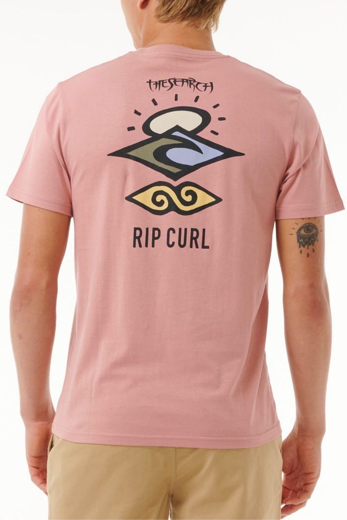 A person wearing a light pink Rip Curl Search Icon Tee, featuring abstract graphic art and "THE SEARCH" text on the back. Made from soft organic cotton, the shirt's unique design is shown from behind.