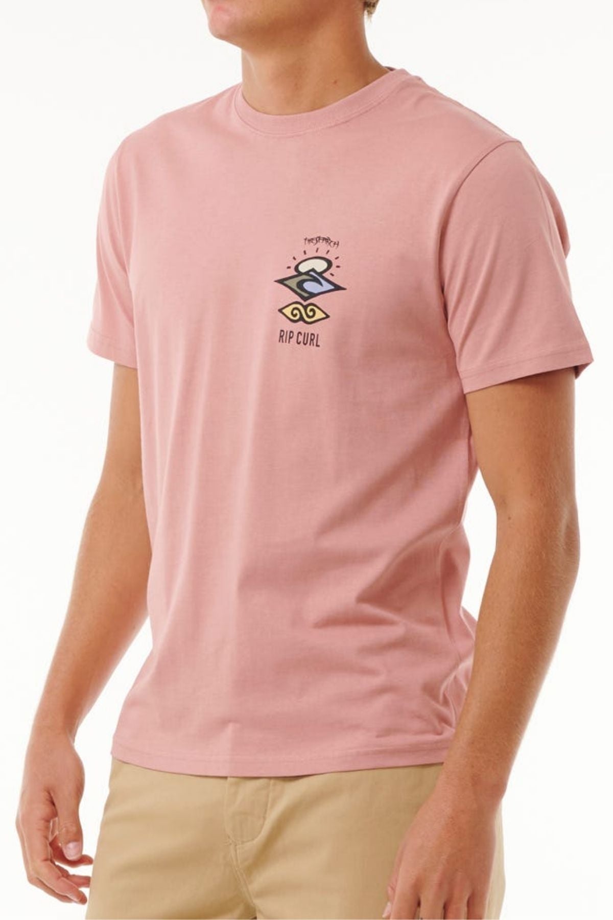 Someone wears a light pink Rip Curl Search Icon Tee made from organic cotton with a small black logo and design on the left chest, paired with beige pants. The background is plain white.