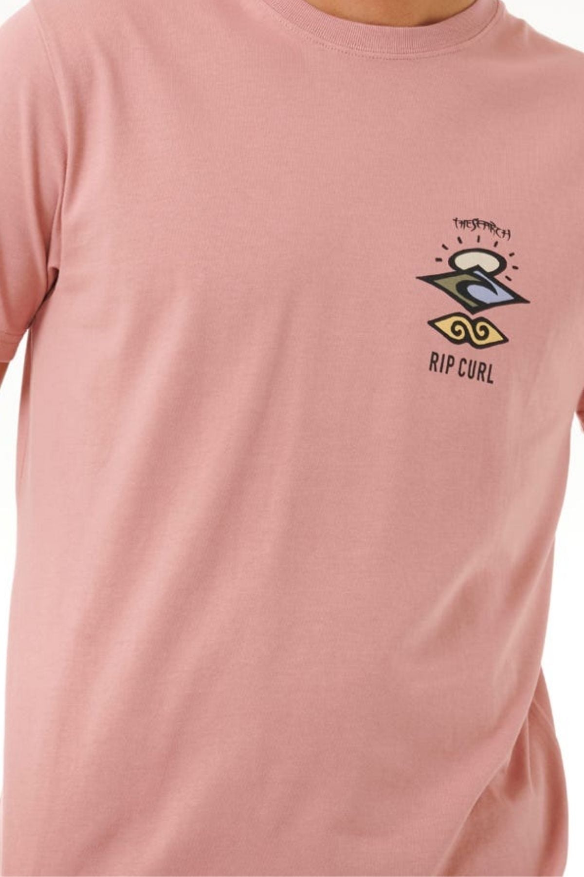 A person wears a light pink Rip Curl Search Icon Tee made from organic cotton, featuring a small colorful logo and design on the left chest. The person's head is not visible in the image.