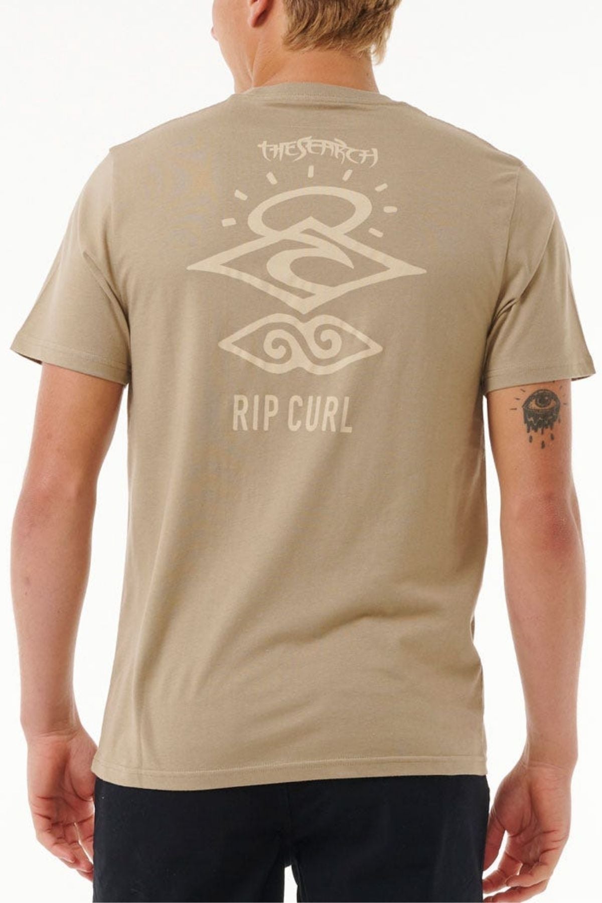 A person sports the Rip Curl Search Icon Tee in beige, featuring abstract designs and "RIP CURL" on the back. The organic cotton tee pairs well with their short, light-colored hair and a small right forearm tattoo against a plain white backdrop.