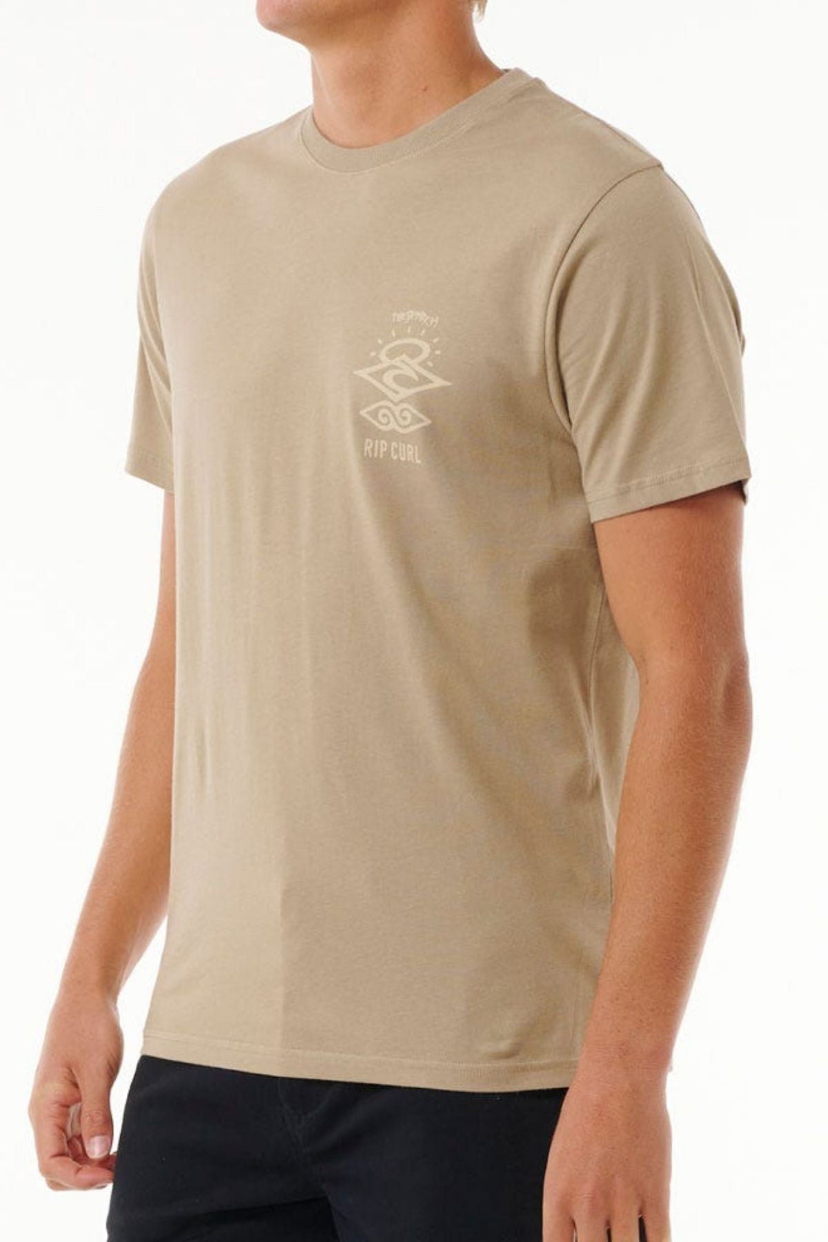 A person is wearing a beige Rip Curl Search Icon Tee, crafted from soft organic cotton with a small logo on the left chest. This short-sleeved crew neck tee is paired with black pants against a neutral background.