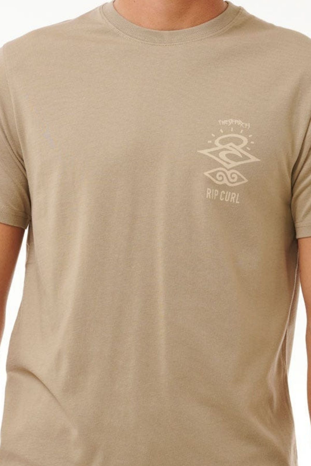 A person wears a beige Relaxed Fit Rip Curl Search Icon Tee, featuring a small design and "RIP CURL" text on the chest. Made from Organic Cotton, it offers comfort and style against a plain white backdrop.