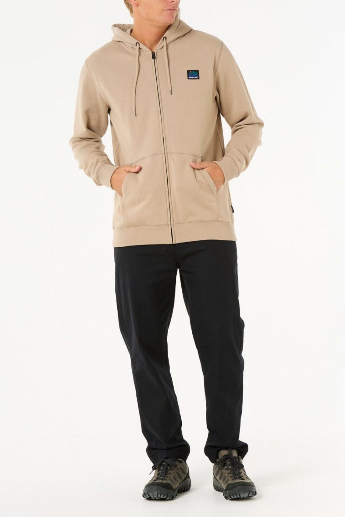 A person wears a beige Rip Curl Search Zip Thru Hood with a small chest logo, metal zipper closure, black pants, and dark athletic shoes, standing with hands in pockets against a plain white background.