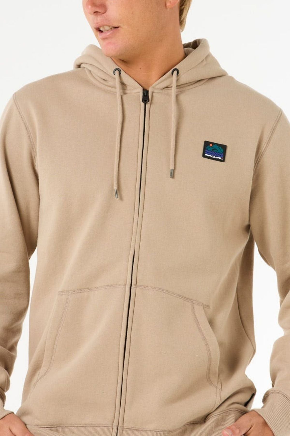 A person wearing the Rip Curl Search Zip Thru Hood in light beige, featuring a small mountain patch on the left chest, metal zipper, drawstrings, and front pockets, is facing slightly to the side.