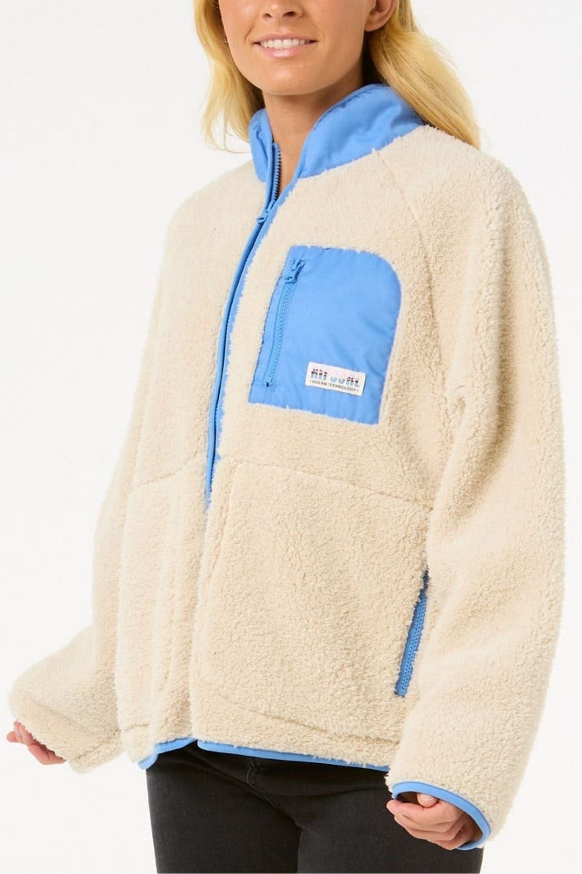 A person wears the Rip Curl Shore Break Polar Fleece, a cream-colored, relaxed fit pullover with light blue chest pocket, collar, and zippers. Made from 100% polyester 300GSM thermal microfleece. Background is plain white.