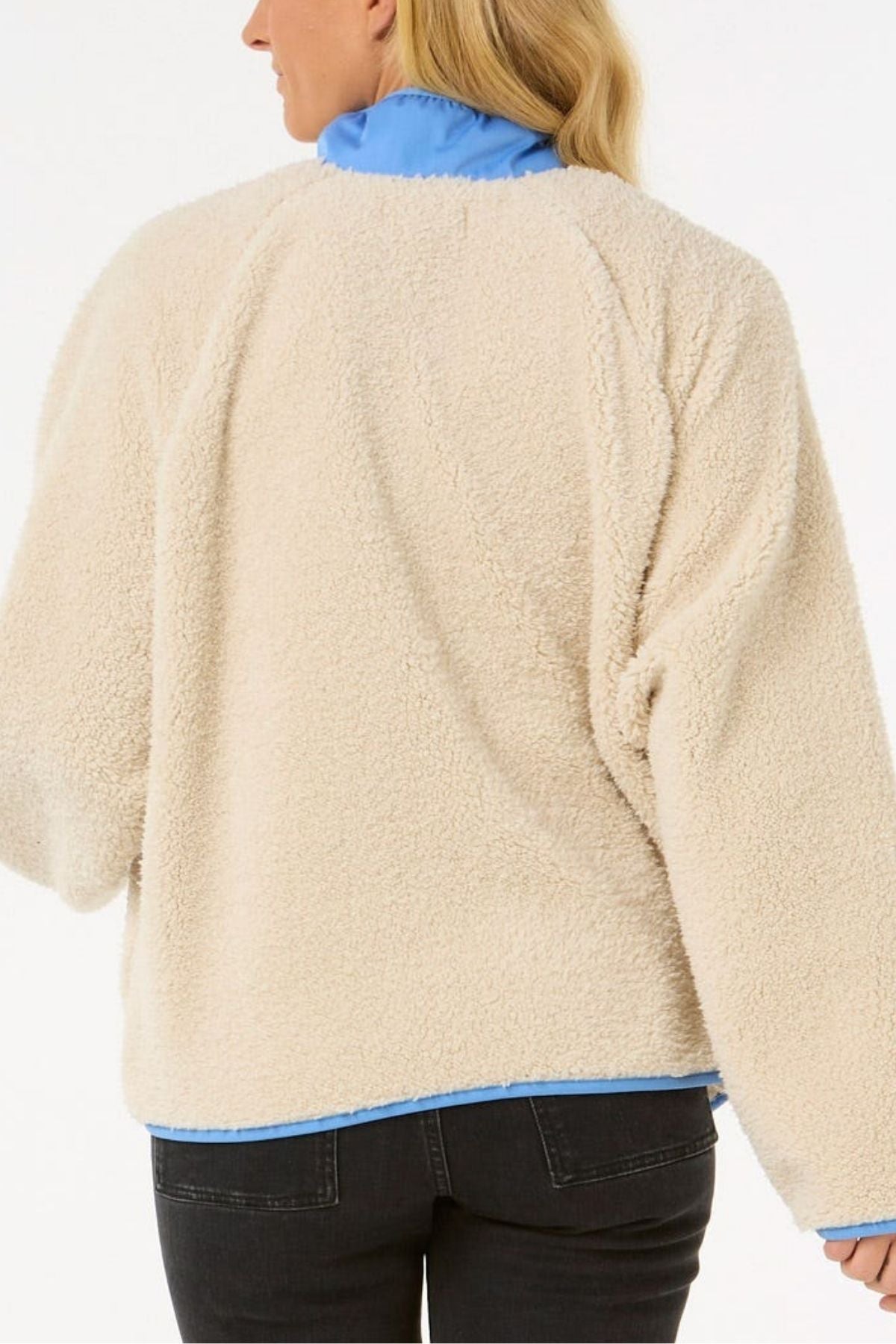 A person with long blonde hair is seen from the back, wearing the Rip Curl Shore Break Polar Fleece in cream over a blue shirt and dark jeans, standing against a plain light background.