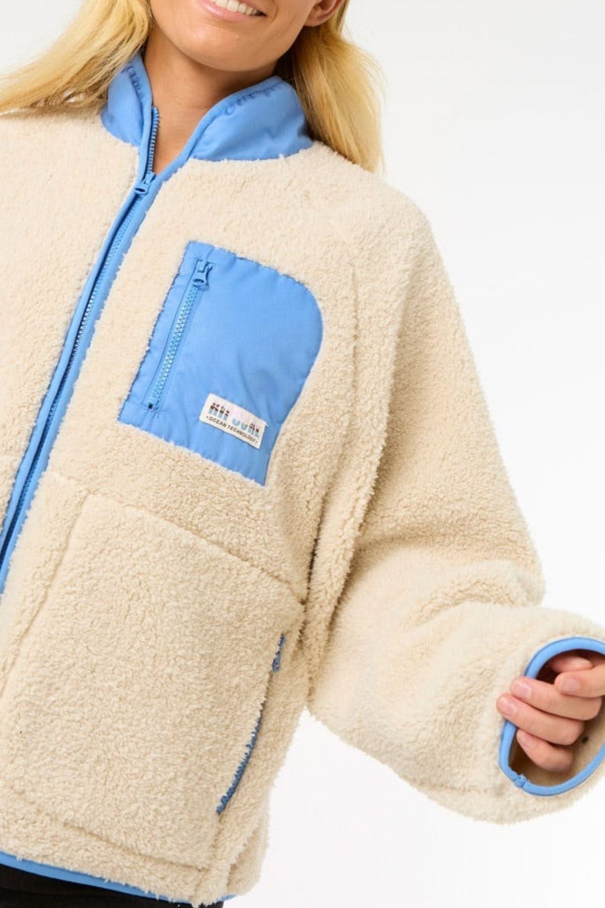 A person wearing the Rip Curl Shore Break Polar Fleece, a cream 100% Polyester 300GSM sherpa jacket with light blue collar, chest pocket, and sleeve accents, smiles against a plain white background.