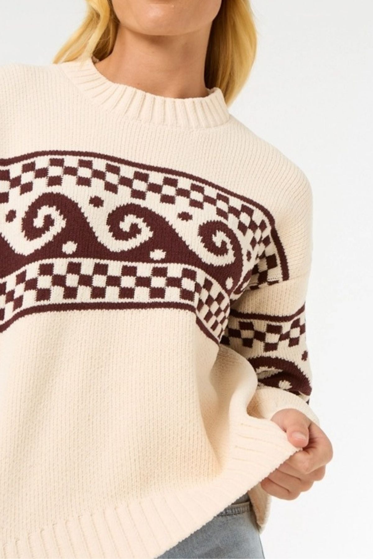 A person wearing the Rip Curl Soleil Knit Crew, an oversized cream-colored sweater with brown geometric and swirl patterns on the chest and sleeves, holds the hem. The person's head is partially out of frame.