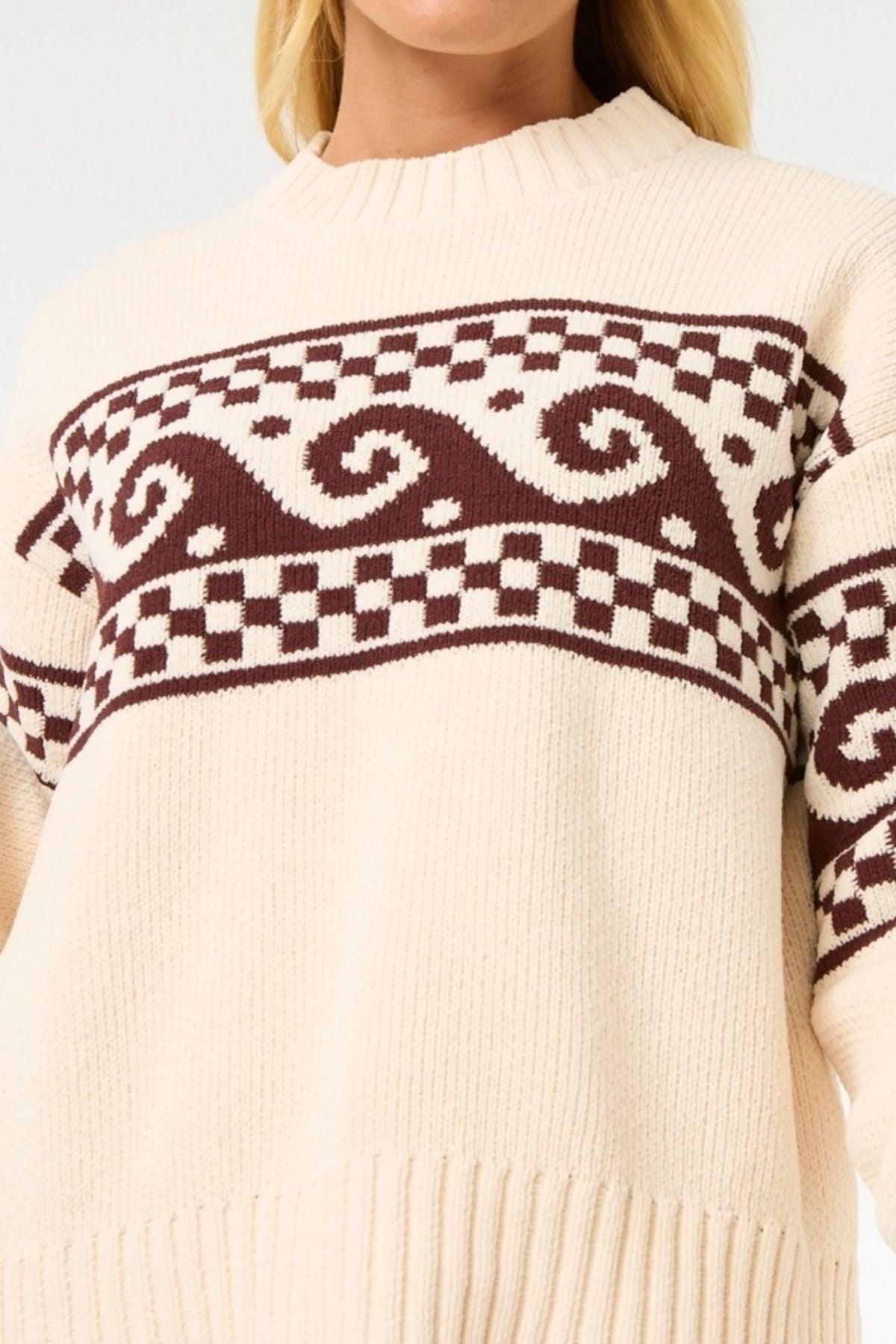 A person wears the Rip Curl Soleil Knit Crew, an oversized cream knit with brown geometric patterns. The image displays their upper body and part of the face.