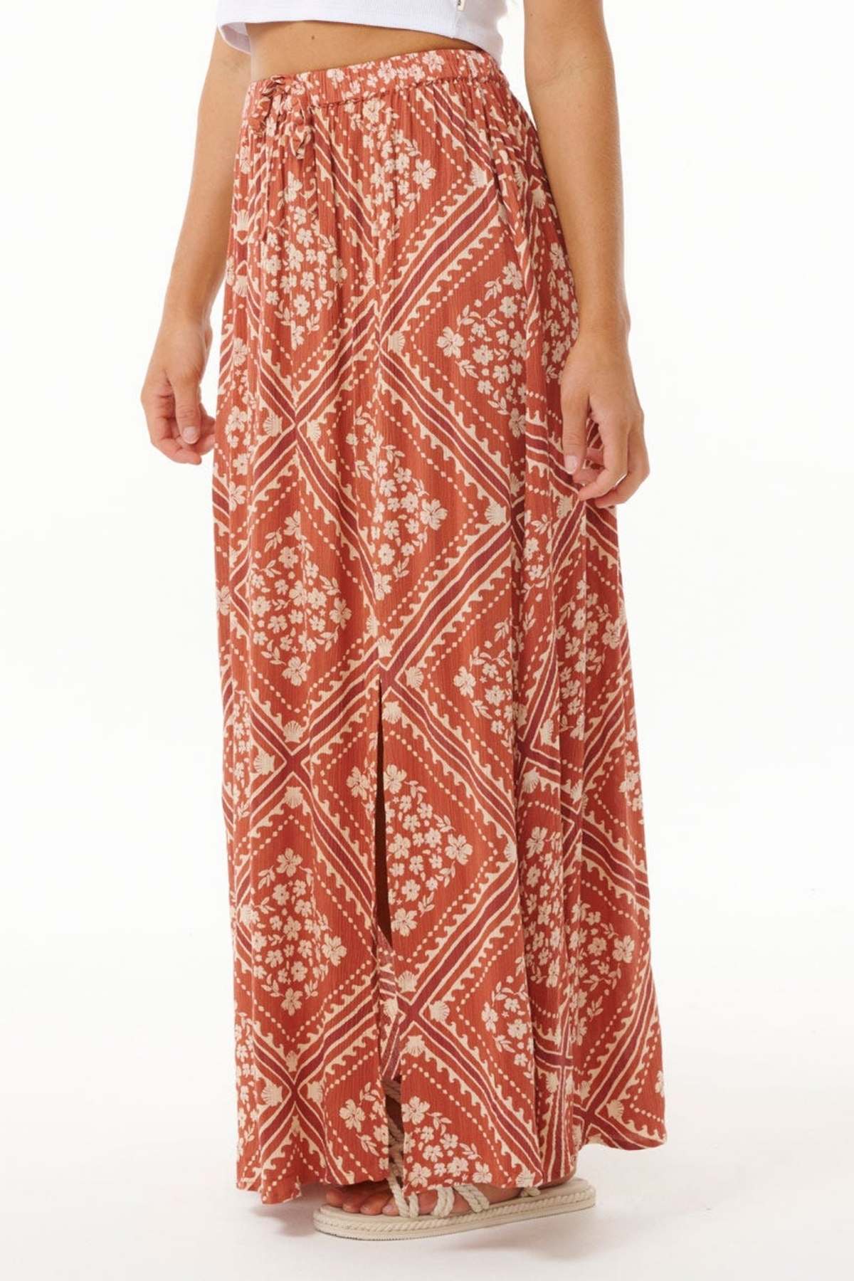 A person sporting the Rip Curl Soleil Maxi Skirt, showcasing its Lenzing Ecovero fabric with a floral and geometric design in rust and white hues, paired elegantly with a white top. This skirt emphasizes sustainable fashion through its relaxed fit and front slit.