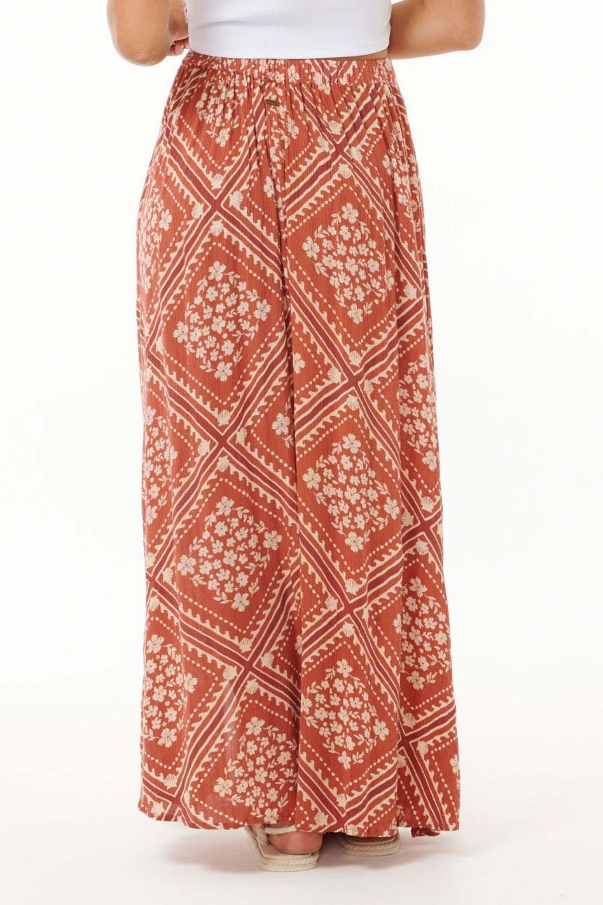 A person is wearing the Rip Curl Soleil Maxi Skirt, a long and flowy piece designed with a red and white geometric and floral pattern made from Lenzing Ecovero. This sustainable fashion skirt includes an elastic waistband and extends to the ankles. The person is standing with their back to the camera on a light background.