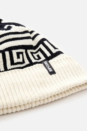 A close-up of the Rip Curl Soleil Regular Pom Pom Beanie, showcasing its black and white geometric intarsia design, folded ribbed brim, and a small fabric tag with the "RIP CURL" brand name attached to the brim.
