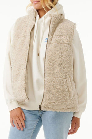 A person wears the Rip Curl Soleil Revo Puffer Vest in light beige over a cream hoodie with visible drawstrings and partial graphic, paired with light blue jeans. The background is plain white.