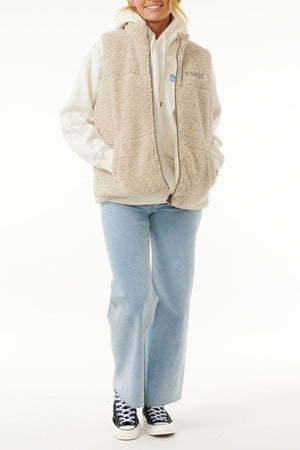 A person wears the Rip Curl Soleil Revo Puffer Vest over a light hoodie, paired with light blue wide-leg jeans and black-and-white sneakers, standing with hands in vest pockets against a white background.