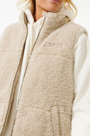 A person wears the cream-colored Rip Curl Soleil Revo Puffer Vest—a fluffy polyester sherpa vest with a zip front, high collar, and subtle chest embroidery—over a white hoodie. Only the lower half of their face is visible.