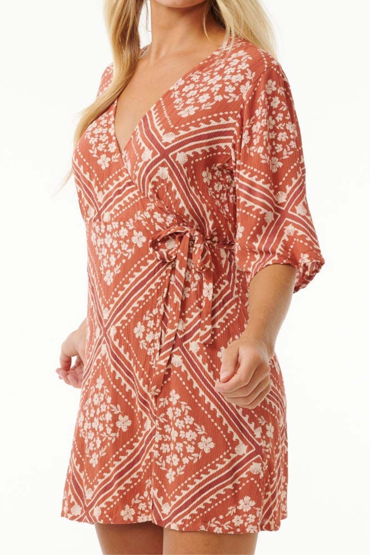 A person dons the Rip Curl Soleil Wrap Mini Dress, showcasing its red and white floral patterns. The dress features a deep V-neckline and three-quarter sleeves. Crafted from Lenzing™ Ecovero™, its eco-friendly fabric stands out against a neutral background, accentuating the dress's design.