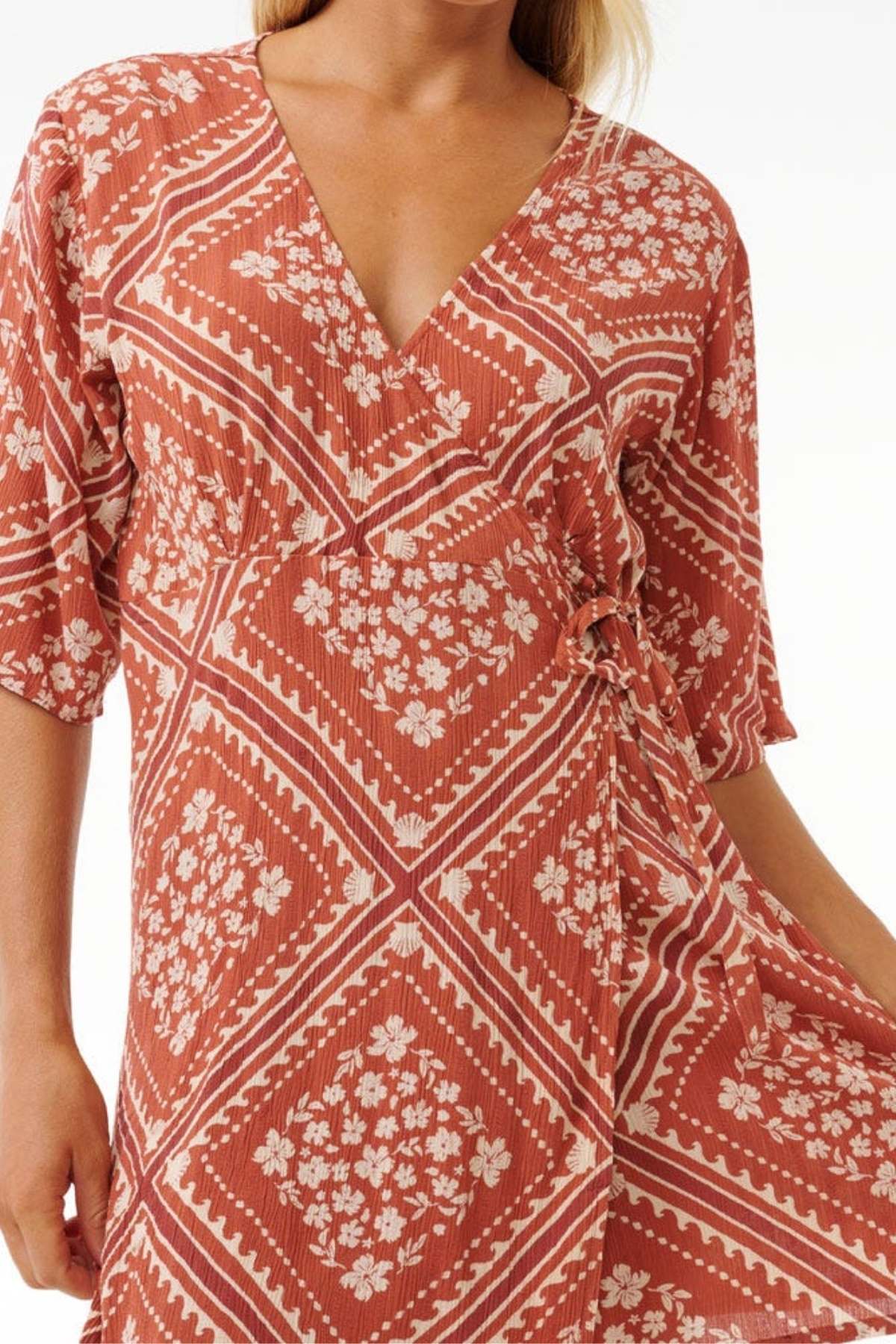 A person wearing the Rip Curl Soleil Wrap Mini Dress in rust, adorned with white floral and geometric patterns. This dress has a V-neckline, short sleeves, and is made from sustainable Lenzing™ Ecovero™ for an eco-friendly appeal.