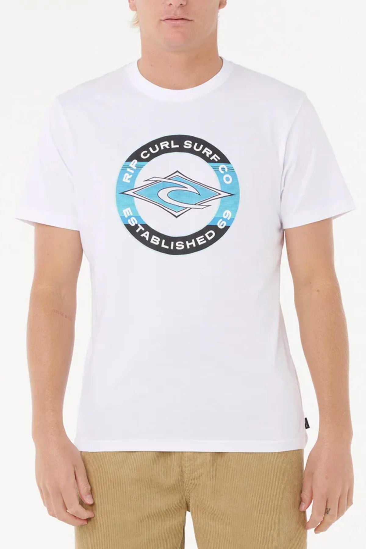 Wearing the Rip Curl Staple Filler Tee in white with a circular blue and black "Rip Curl Surf Co Established 69" chest logo and tan corduroy pants, a person stands against a plain white background.