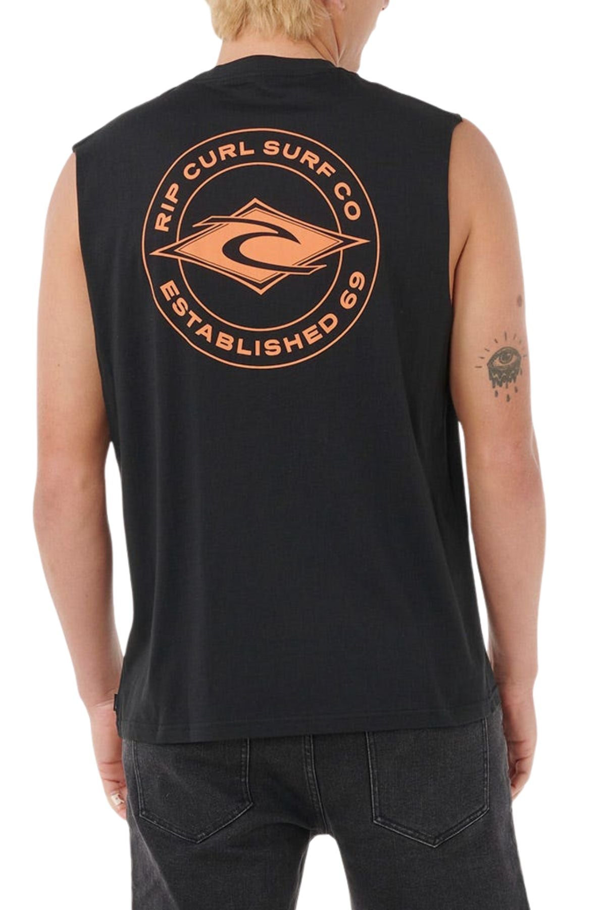 A person with short blond hair wears a black Rip Curl Staple Muscle shirt made from 100% cotton, featuring an orange "Rip Curl Surf Co Established 69" logo on the back and has a tattoo of an eye with rays on their left arm.