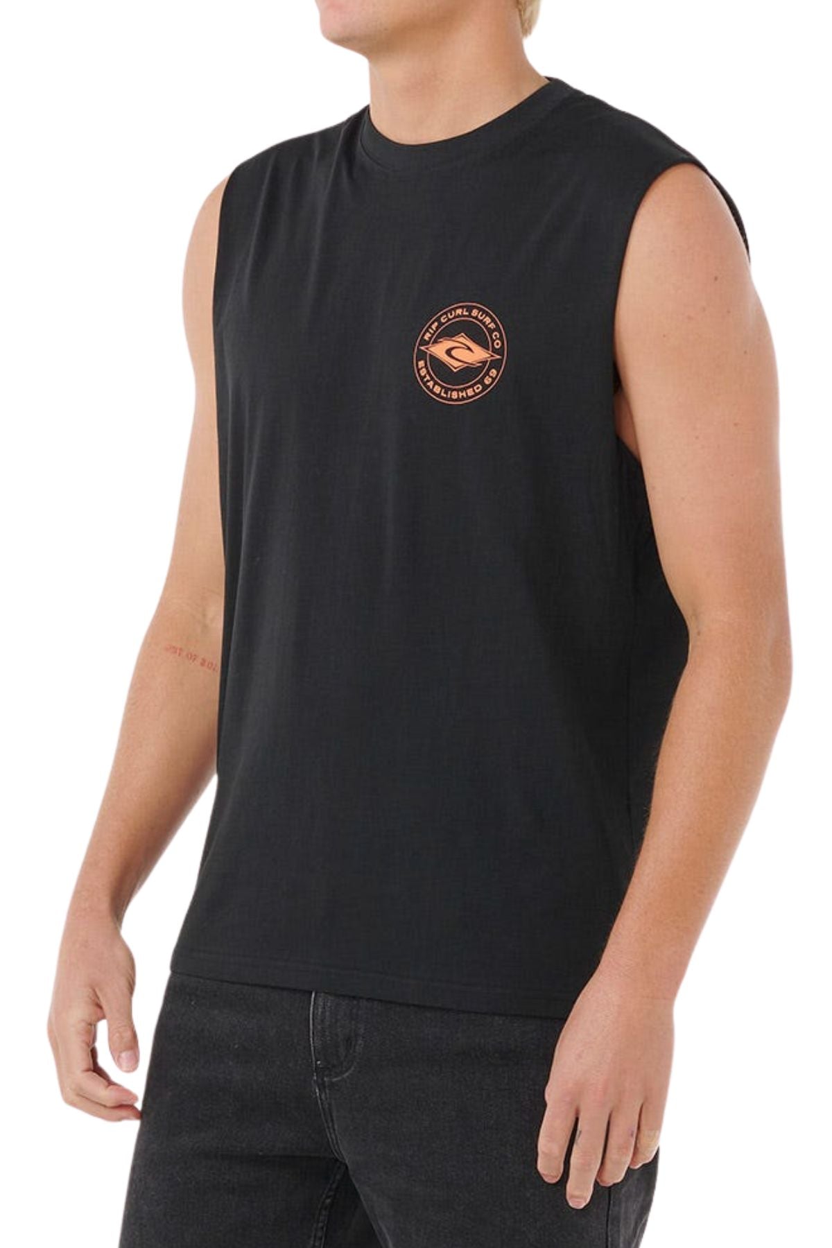 A person wears the Rip Curl Staple Muscle, a sleeveless black 100% cotton shirt with a small orange circular logo on the left chest, paired with dark pants, standing against a plain white background.