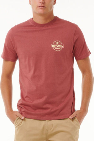 A person in a stylish Rip Curl Staple Tee, featuring a small logo on the chest of its red cotton jersey against a plain white background, stands with hands in the pockets of their beige pants.