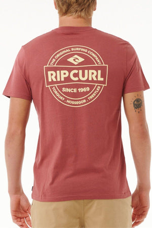 A person wears a maroon Rip Curl Staple Tee made from cotton jersey, featuring a large circular logo on the back that says "RIP CURL SINCE 1969," highlighting "TORQUAY, HOSSEGOR, TRESTLES," complemented by a small wrist tattoo.