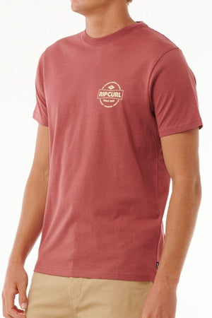 Wearing a stylish maroon Rip Curl Staple Tee, short-sleeved and crafted from soft cotton jersey with a logo on the chest, paired with beige pants, a person poses effortlessly against a plain white background.