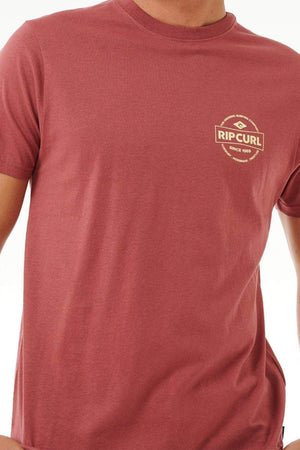 A person is wearing a maroon Rip Curl Staple Tee with a circular logo on the left chest. This stylish short-sleeve tee is crafted from soft cotton jersey against a plain white background.