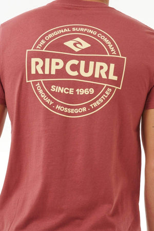 Sporting the Rip Curl Staple Tee, a person presents a casual vibe with its stylish red design, featuring the "Rip Curl" logo and "The Original Surfing Company, Since 1969, Torquay, Hossegor, Trestles" in a circular pattern on the back.