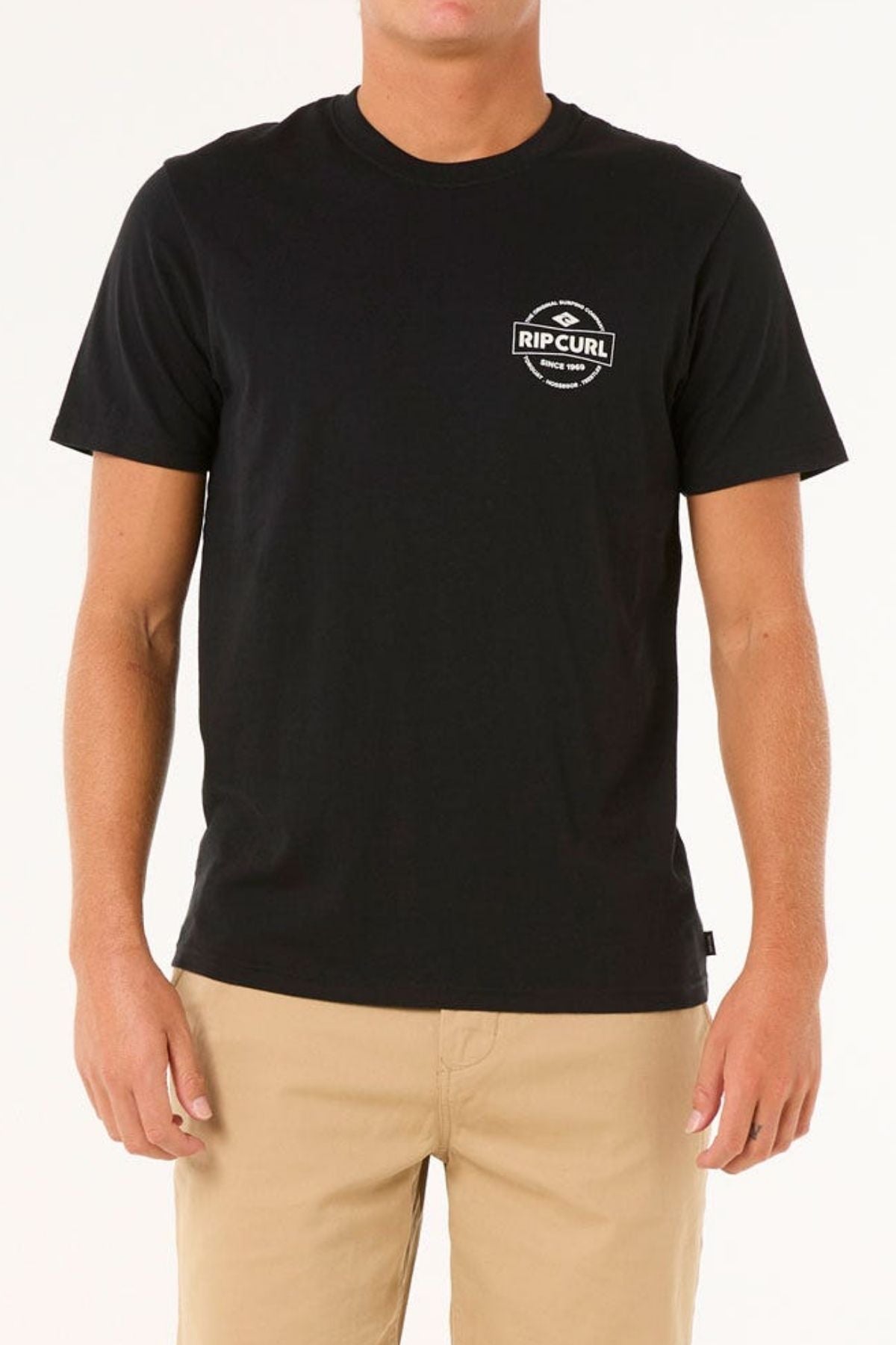 Wearing a black Rip Curl Staple Tee, crafted from soft cotton jersey and featuring a small chest logo, a person stands against a plain background. This simply designed short-sleeve T-shirt is stylishly paired with beige pants, ideal for casual occasions.