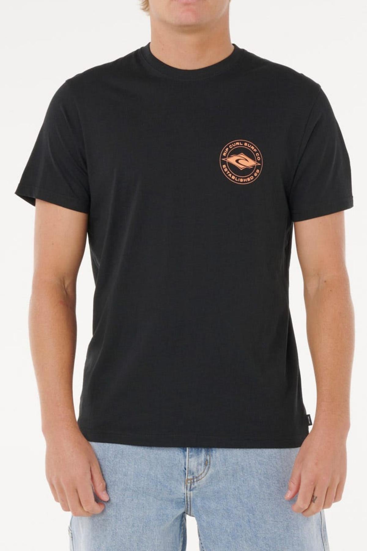Wearing the Rip Curl Staple Tee—plain black cotton with a small circular orange logo on the left chest—and light blue jeans, a person stands against a white background. The tee delivers classic style and comfort.