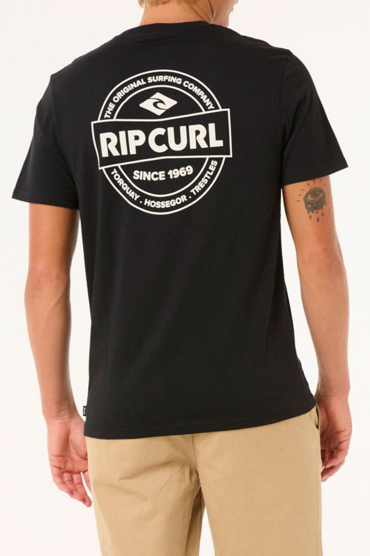 A person wears a black Rip Curl Staple Tee made from soft cotton jersey with a large logo and "Since 1969: Torquay, Hossegor, Trestles" text on the back. They're also in beige pants with a small tattoo on their arm—ideal for any casual occasion.
