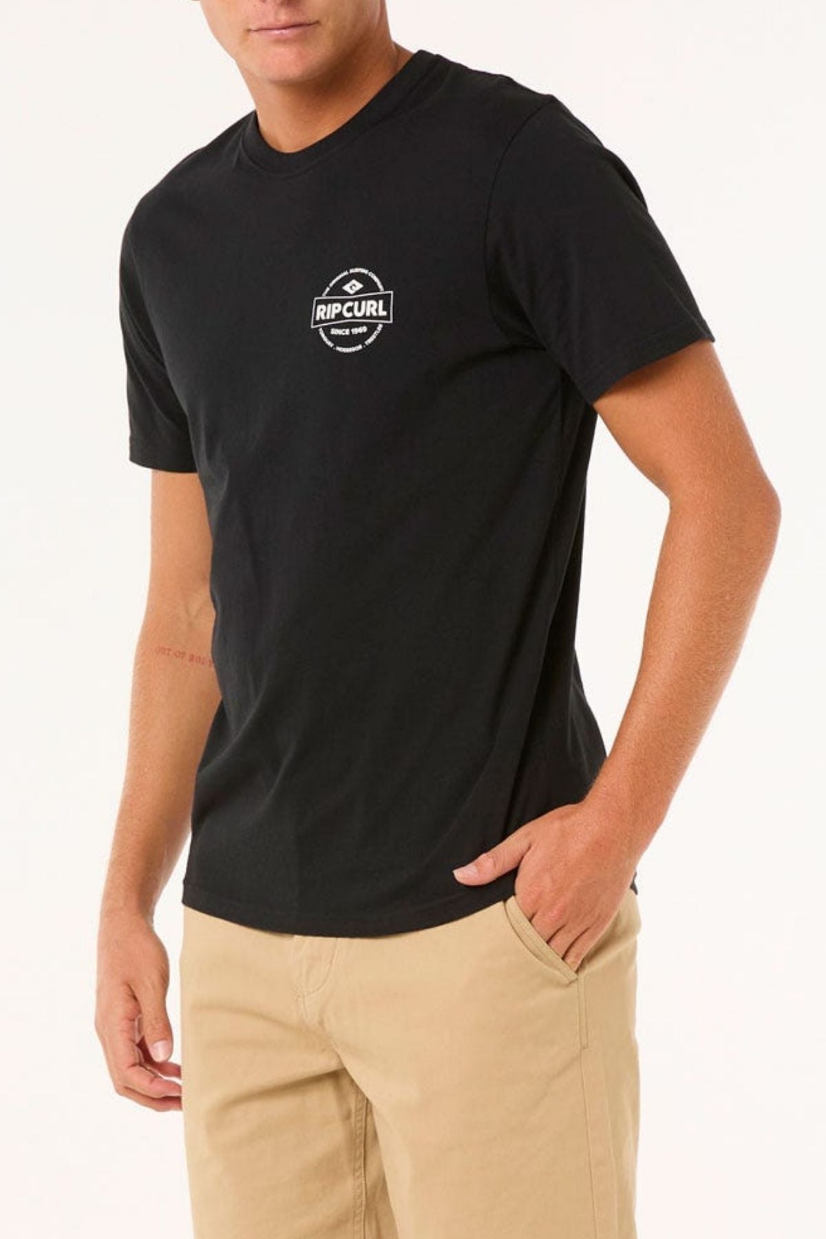 Wearing a black Rip Curl Staple Tee made of soft cotton jersey, a man pairs it with beige pants, standing against a white background with one hand in his pocket, showcasing laid-back style for casual occasions.