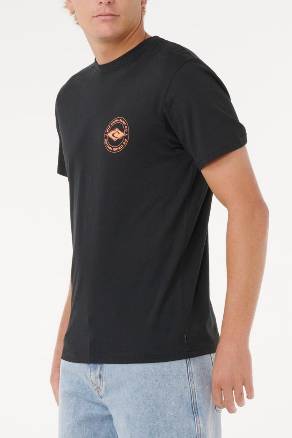 A person wears the Rip Curl Staple Tee in black, featuring a small round logo on the left chest, paired with light blue jeans and standing against a white background. This cotton tee provides a comfortable everyday fit.