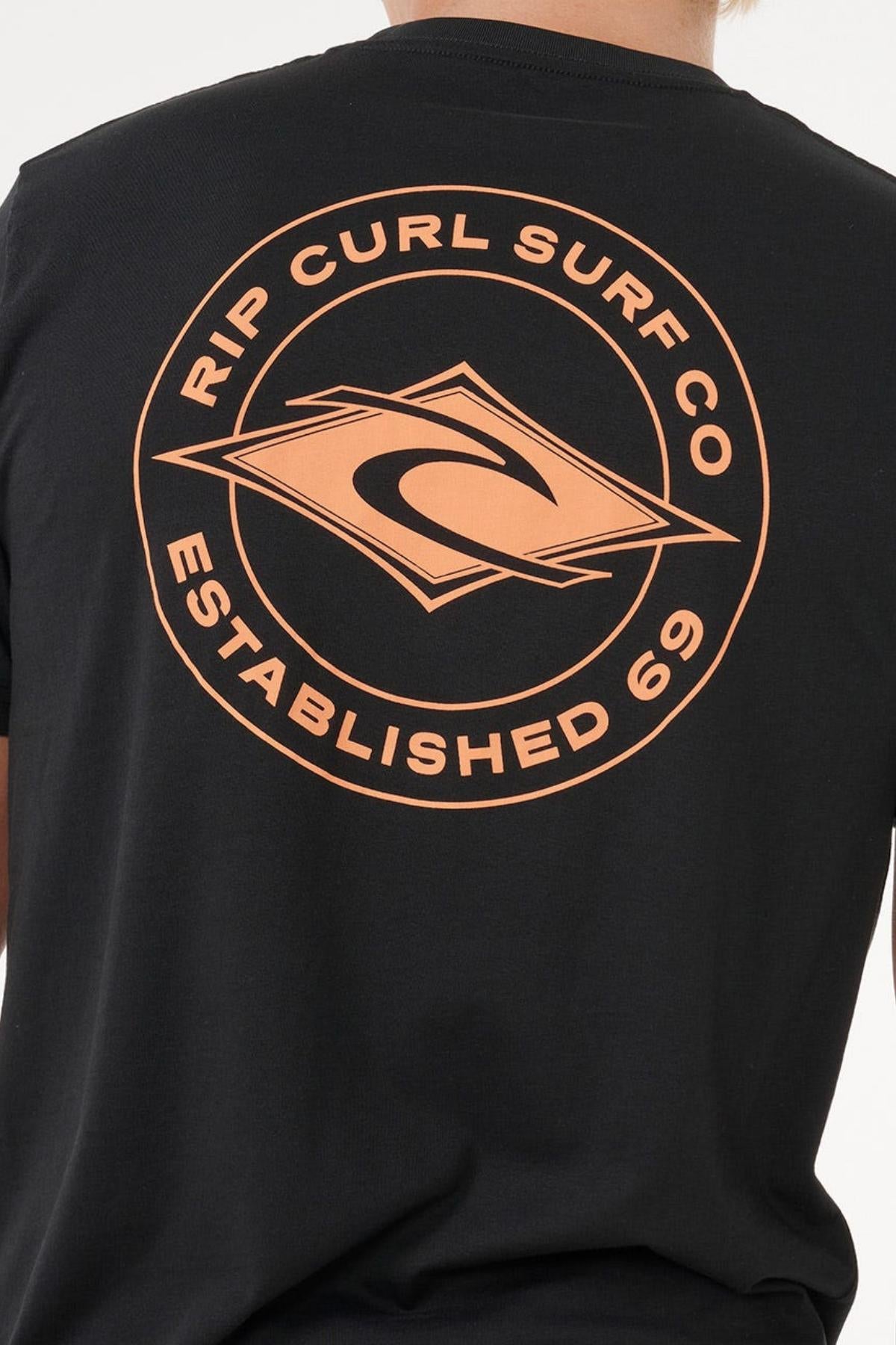 A person wears the Rip Curl Staple Tee, a black cotton t-shirt with an orange circular "RIP CURL SURF CO ESTABLISHED 69" logo and diamond wave emblem on the back for a comfortable fit.