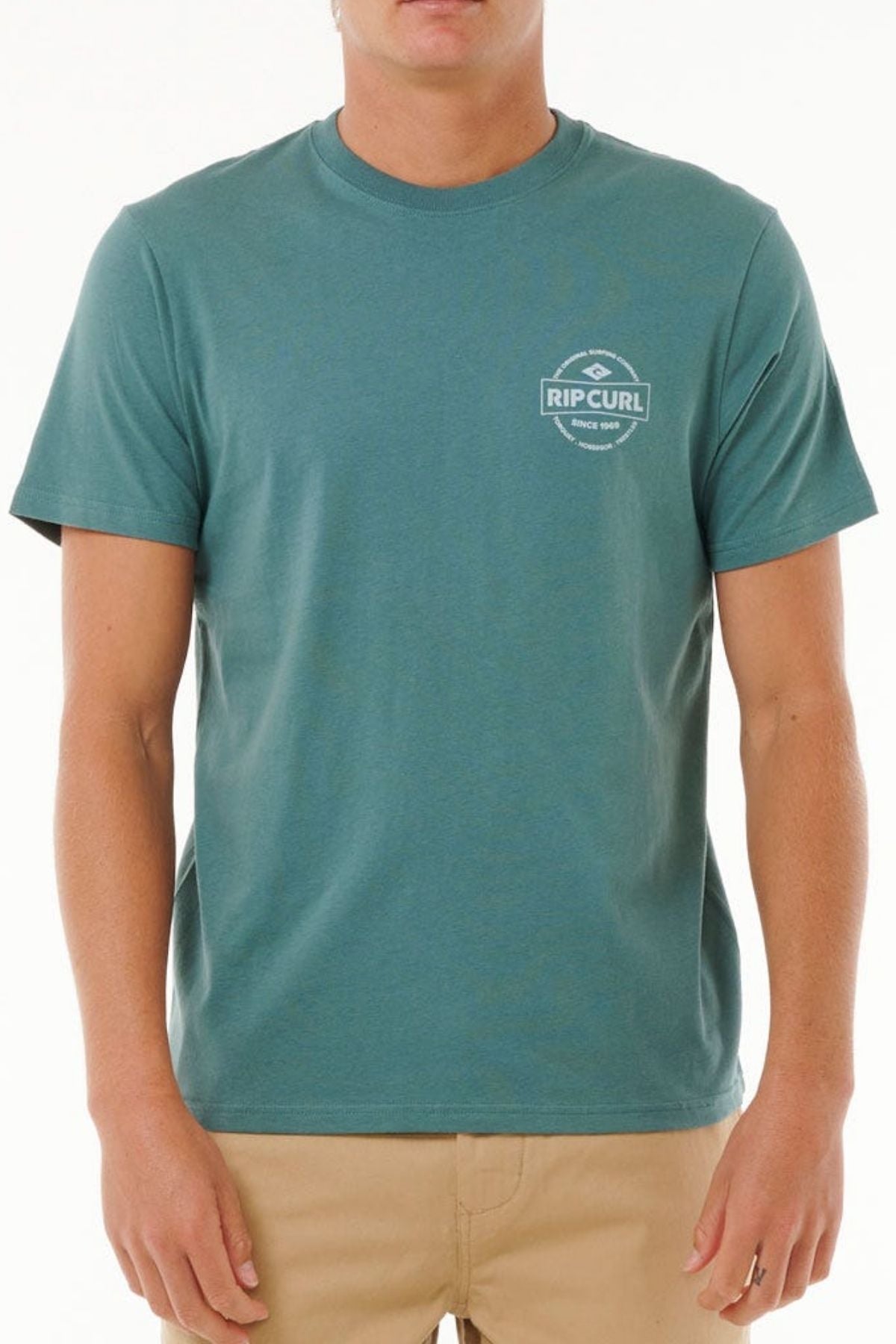 Wearing the Rip Curl Staple Tee in teal, made from soft cotton jersey with a small white logo on the left chest, and beige pants, a person stands against a plain white background.