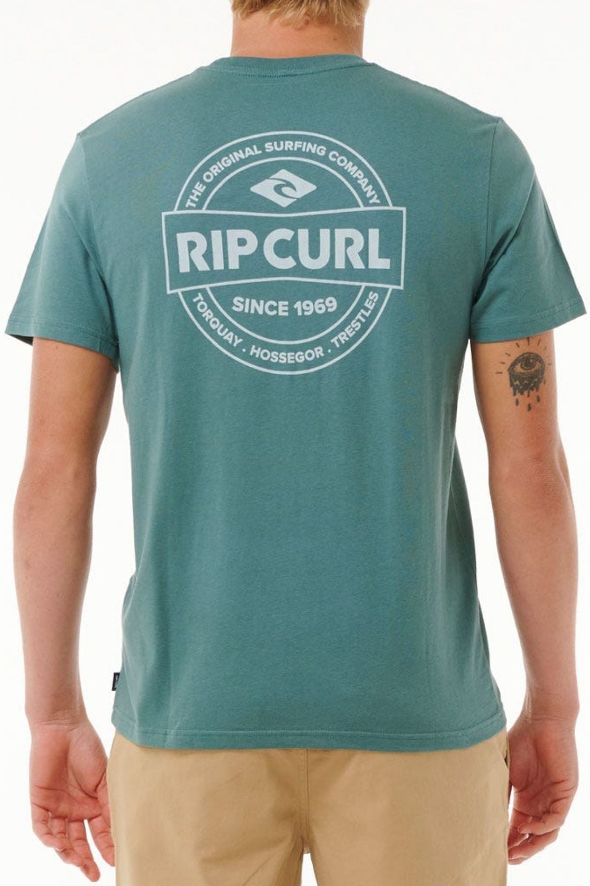 Wearing a teal Rip Curl Staple Tee in soft cotton jersey with a bold white "Rip Curl Since 1969" logo and beach locations on the back, a person faces away, revealing a tattoo on their left arm.